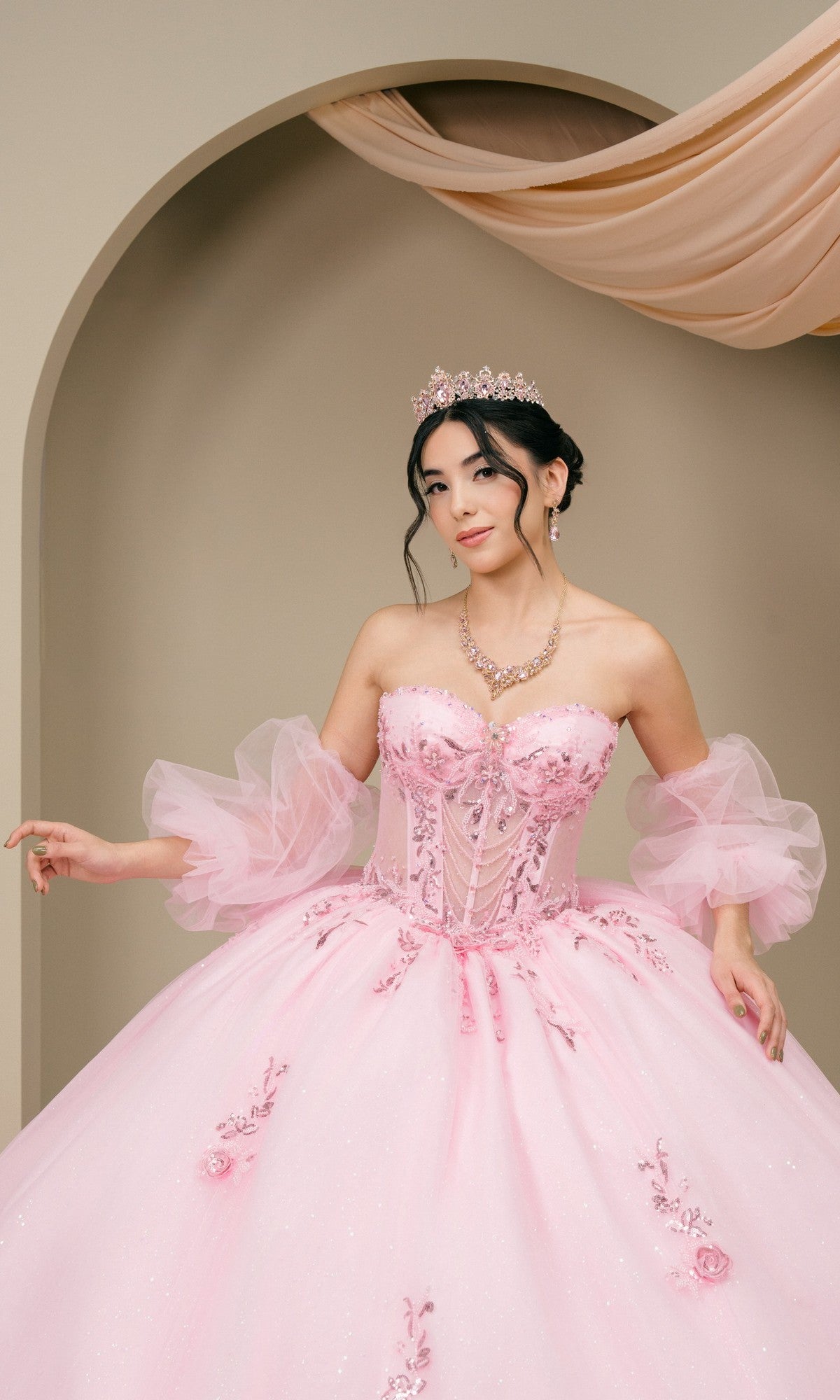 A woman in the Quinceanera Dress 8380J by Cinderella Couture, featuring pink floral details and sheer puffy sleeves, stands before an arched backdrop with draped fabric, accessorized with a tiara and jewelry.